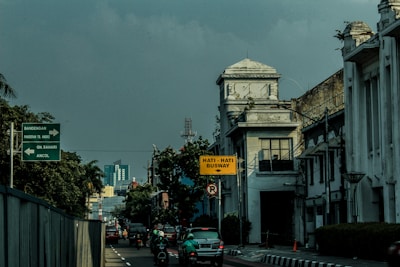 A street scene featuring an urban environment with historical architecture on the right side, including a building with a clock tower. Several vehicles and motorbikes are on the road, and traffic signs in Indonesian are visible, including one cautioning about a busway.