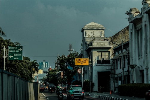 A street scene featuring an urban environment with historical architecture on the right side, including a building with a clock tower. Several vehicles and motorbikes are on the road, and traffic signs in Indonesian are visible, including one cautioning about a busway.