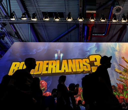 Silhouettes of people walk past a large promotional display for the game 'Borderlands 3', featuring vibrant artwork with roses and gears. The environment is dimly lit with overhead lights and industrial elements visible in the background, creating a dramatic contrast with the colorful poster.