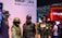Four individuals dressed in tactical gear stand in front of a display for 'Rainbow Six Siege' and a hashtag '#UBIGAMESCOM'. One wears an FBI cap and sunglasses, while another has face paint resembling a skull. The environment appears to be a gaming or cosplay event with colorful lights in the background.