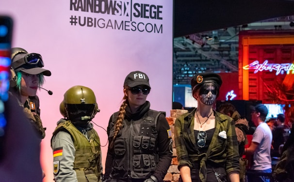 Four individuals dressed in tactical gear stand in front of a display for 'Rainbow Six Siege' and a hashtag '#UBIGAMESCOM'. One wears an FBI cap and sunglasses, while another has face paint resembling a skull. The environment appears to be a gaming or cosplay event with colorful lights in the background.