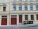 A residential building with beige walls and multiple red-framed windows, featuring ornate architectural details. There are two red garage doors with an additional red front door between them. On the right side, several black and yellow garbage bins are lined up along the sidewalk.