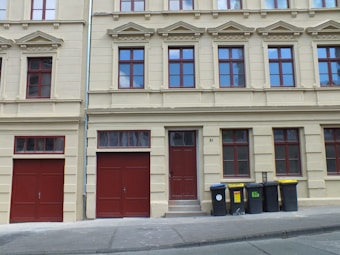 A residential building with beige walls and multiple red-framed windows, featuring ornate architectural details. There are two red garage doors with an additional red front door between them. On the right side, several black and yellow garbage bins are lined up along the sidewalk.