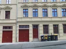 A residential building with beige walls and multiple red-framed windows, featuring ornate architectural details. There are two red garage doors with an additional red front door between them. On the right side, several black and yellow garbage bins are lined up along the sidewalk.