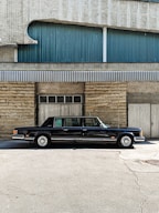 A classic black limousine is parked in front of a building with a textured facade. The building features a combination of stone and corrugated metal panels, with blue and grey tones. The image is taken on a sunny day, casting shadows on the ground.
