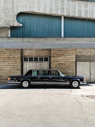 A classic black limousine is parked in front of a building with a textured facade. The building features a combination of stone and corrugated metal panels, with blue and grey tones. The image is taken on a sunny day, casting shadows on the ground.