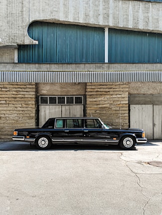 A classic black limousine is parked in front of a building with a textured facade. The building features a combination of stone and corrugated metal panels, with blue and grey tones. The image is taken on a sunny day, casting shadows on the ground.