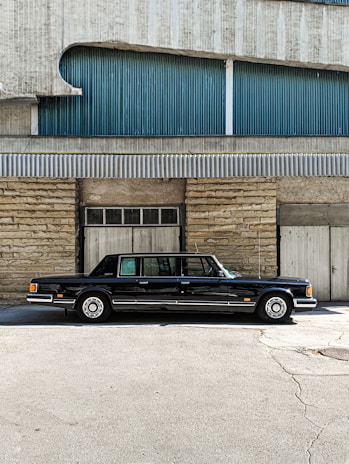 A classic black limousine is parked in front of a building with a textured facade. The building features a combination of stone and corrugated metal panels, with blue and grey tones. The image is taken on a sunny day, casting shadows on the ground.