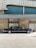 A classic black limousine is parked in front of a building with a textured facade. The building features a combination of stone and corrugated metal panels, with blue and grey tones. The image is taken on a sunny day, casting shadows on the ground.
