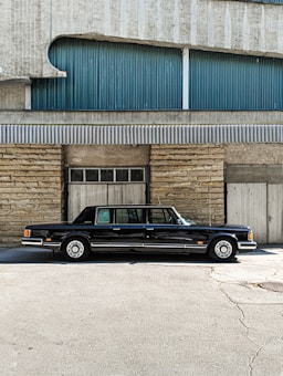 A classic black limousine is parked in front of a building with a textured facade. The building features a combination of stone and corrugated metal panels, with blue and grey tones. The image is taken on a sunny day, casting shadows on the ground.