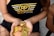 A person holds a collection of shiny, gold and copper-colored cryptocurrency tokens in their hands. They are wearing a black tank top with a logo that reads 'RP Real Poker' in bold letters. The person is also in fishnet stockings and shorts, sitting beside a textured bag.