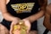 A person holds a collection of shiny, gold and copper-colored cryptocurrency tokens in their hands. They are wearing a black tank top with a logo that reads 'RP Real Poker' in bold letters. The person is also in fishnet stockings and shorts, sitting beside a textured bag.