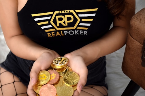 A person holds a collection of shiny, gold and copper-colored cryptocurrency tokens in their hands. They are wearing a black tank top with a logo that reads 'RP Real Poker' in bold letters. The person is also in fishnet stockings and shorts, sitting beside a textured bag.