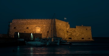 An ancient stone fortress stands illuminated by soft yellow lights against a dark night sky. The structure is massive, with sturdy walls and battlements visible. Below it, several small boats are docked peacefully along the waterfront. The Greek flag is seen waving atop the fort, adding a cultural touch.