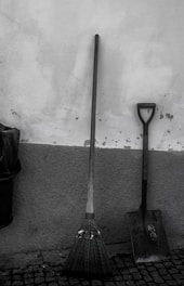 A broom and a shovel are leaning against a textured wall. The broom features green stripes near the bristles, and both tools stand on a cobblestone floor. A trash bin is partially visible on the left.