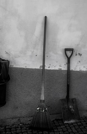 A broom and a shovel are leaning against a textured wall. The broom features green stripes near the bristles, and both tools stand on a cobblestone floor. A trash bin is partially visible on the left.