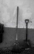 A broom and a shovel are leaning against a textured wall. The broom features green stripes near the bristles, and both tools stand on a cobblestone floor. A trash bin is partially visible on the left.