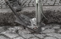A person stands on a cobblestone surface holding a broom. Next to them, a shovel is filled with crumpled paper, suggesting street cleaning. The scene is focused on the lower part of the person's legs and feet, and the tools used in the cleaning process.