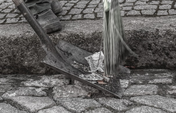 A person stands on a cobblestone surface holding a broom. Next to them, a shovel is filled with crumpled paper, suggesting street cleaning. The scene is focused on the lower part of the person's legs and feet, and the tools used in the cleaning process.