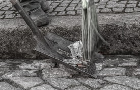 A person stands on a cobblestone surface holding a broom. Next to them, a shovel is filled with crumpled paper, suggesting street cleaning. The scene is focused on the lower part of the person's legs and feet, and the tools used in the cleaning process.