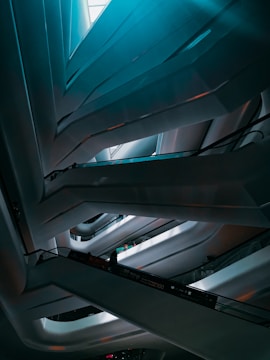 A modern interior featuring abstract, sweeping architectural lines that form a multi-level structure. The lighting is subtle, casting soft shadows and highlights across the sleek surfaces. An escalator can be seen with a few silhouetted figures standing on it, adding a sense of scale and movement.