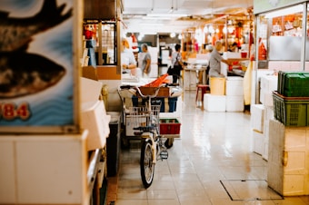 A vibrant indoor market scene features several people busy with various tasks. A bicycle with a basket is placed near a shopping cart filled with items. Stacks of boxes and containers are organized around the market, while posters and signs are visible on the walls. The atmosphere is lively, and the floor reflects the bright lighting.