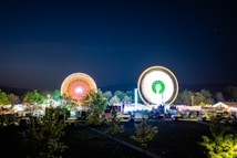 A vibrant fairground at night, featuring two brightly lit Ferris wheels in motion, creating circular light trails. The scene includes illuminated tents and trees in the foreground, with a dark, clear sky above.