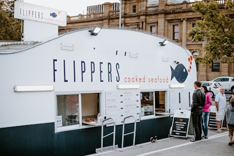 A food stall named Flippers Cooked Seafood is situated outdoors with people standing in front of it. The stall has a clean and minimalist design with a large sign displaying its name and a graphic of a fish. A menu board is visible, offering various seafood dishes. The surrounding area includes some trees and another building in the background.
