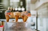 Three ice cream cones with different flavors are arranged side by side in a metal holder. The first cone features vibrant swirls of blue and red ice cream, the second has a creamy vanilla with chocolate chips, and the third appears to have a similar color to the first, possibly with additional flavors. A blurred background suggests an interior of an ice cream shop, with a menu slightly visible.