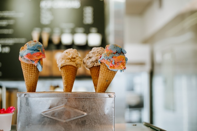 Three ice cream cones with different flavors are arranged side by side in a metal holder. The first cone features vibrant swirls of blue and red ice cream, the second has a creamy vanilla with chocolate chips, and the third appears to have a similar color to the first, possibly with additional flavors. A blurred background suggests an interior of an ice cream shop, with a menu slightly visible.