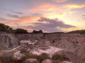 A quarry surrounded by rocky terrain under a colorful sky at sunset, with hues of pink, orange, and purple complementing the scene. Buildings and equipment are visible in the foreground, set against the dramatic landscape.