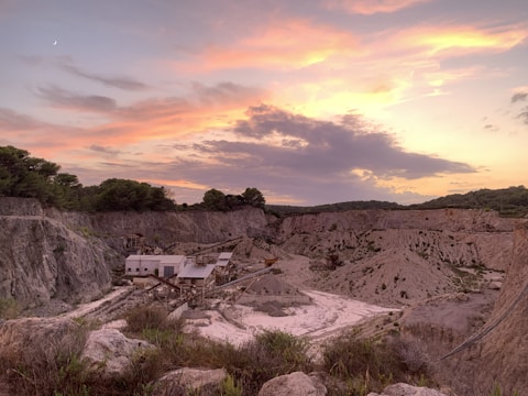 A quarry surrounded by rocky terrain under a colorful sky at sunset, with hues of pink, orange, and purple complementing the scene. Buildings and equipment are visible in the foreground, set against the dramatic landscape.