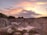 A quarry surrounded by rocky terrain under a colorful sky at sunset, with hues of pink, orange, and purple complementing the scene. Buildings and equipment are visible in the foreground, set against the dramatic landscape.