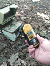A hand is holding a Geiger counter displaying a reading of 12.9 µSv/h. On the ground, among dry leaves, there is a small, worn-out metal container with coins on top. The setting appears to be outdoors, possibly in a forest or park area, as indicated by the leaves and natural debris.