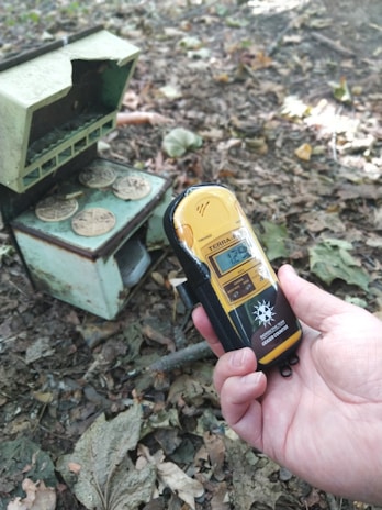 A hand is holding a Geiger counter displaying a reading of 12.9 µSv/h. On the ground, among dry leaves, there is a small, worn-out metal container with coins on top. The setting appears to be outdoors, possibly in a forest or park area, as indicated by the leaves and natural debris.