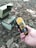 A hand is holding a Geiger counter displaying a reading of 12.9 µSv/h. On the ground, among dry leaves, there is a small, worn-out metal container with coins on top. The setting appears to be outdoors, possibly in a forest or park area, as indicated by the leaves and natural debris.