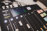 A professional audio mixing console with various controls and buttons is visible. The display screen shows audio levels, and the panel includes sliders, buttons, and knobs lit with different colored lights.