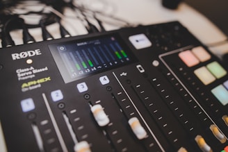 A professional audio mixing console with various controls and buttons is visible. The display screen shows audio levels, and the panel includes sliders, buttons, and knobs lit with different colored lights.