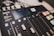 A professional audio mixing console with various controls and buttons is visible. The display screen shows audio levels, and the panel includes sliders, buttons, and knobs lit with different colored lights.