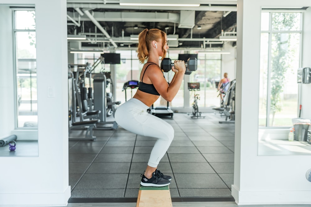 mulher vestindo sutiã esportivo preto e legging branco levantando halteres