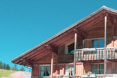 A wooden alpine-style house with a sloped roof and a large balcony. The house features multiple windows and floral decorations on the balcony. The sky is clear, with a vibrant blue hue, and there are some trees visible in the background.