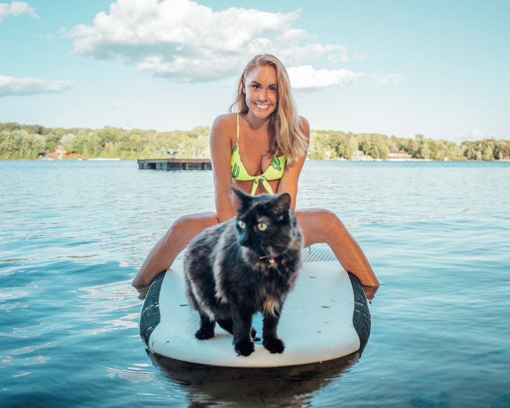 woman wearing green bikini top sitting on black and white surfboard near tortoiseshell cat on blue sea under blue and white skies during daytime