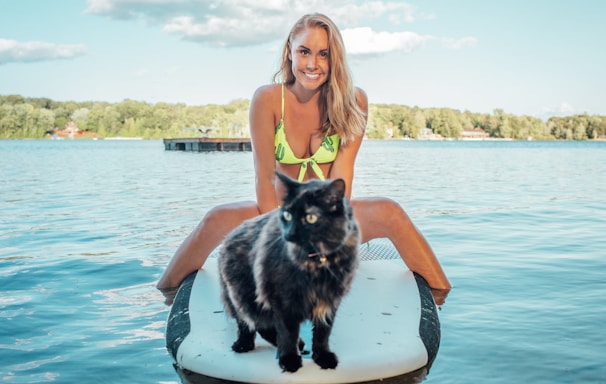 woman wearing green bikini top sitting on black and white surfboard near tortoiseshell cat on blue sea under blue and white skies during daytime
