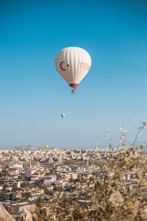 A hot air balloon with Turkish symbols floats above a scenic landscape, featuring a town with numerous buildings and unique rock formations in the background. The clear blue sky adds a sense of openness, while a few other balloons are visible in the distance.