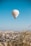 A hot air balloon with Turkish symbols floats above a scenic landscape, featuring a town with numerous buildings and unique rock formations in the background. The clear blue sky adds a sense of openness, while a few other balloons are visible in the distance.