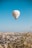 A hot air balloon with Turkish symbols floats above a scenic landscape, featuring a town with numerous buildings and unique rock formations in the background. The clear blue sky adds a sense of openness, while a few other balloons are visible in the distance.