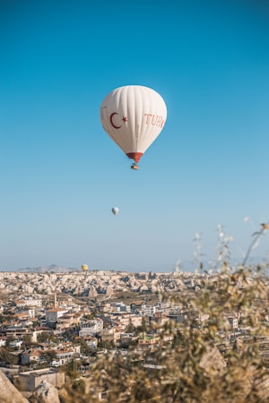 A hot air balloon with Turkish symbols floats above a scenic landscape, featuring a town with numerous buildings and unique rock formations in the background. The clear blue sky adds a sense of openness, while a few other balloons are visible in the distance.