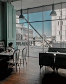 A person sits alone at a table in a modern cafe, working on a laptop. The large window panes reveal an urban scene outside with buildings and a clear blue sky. Natural light floods the room, creating a serene and focused atmosphere.