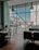 A person sits alone at a table in a modern cafe, working on a laptop. The large window panes reveal an urban scene outside with buildings and a clear blue sky. Natural light floods the room, creating a serene and focused atmosphere.