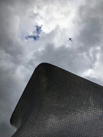 A modern architectural structure with a hexagonal pattern is positioned against a cloudy sky backdrop. An airplane is seen flying high above, creating a dynamic contrast between architecture and sky.
